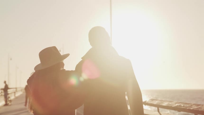 A man and woman walk down a pier with their arms around one another as others walk on the pier with their bikes