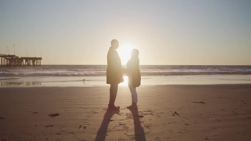 An elderly couple stands together on a sandy beach with the setting sun behind them