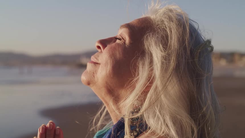 An elderly woman prays while the sun shines on her smiling face on the beach