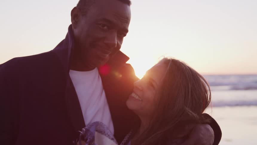 A man and woman smile at one another and hug on the beach