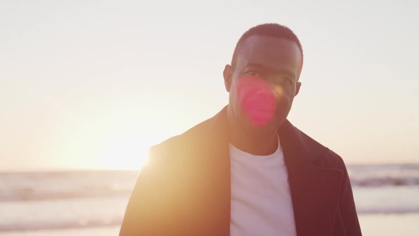 Black man with a black jacket stands and looks at the camera on beach at sunset next to the ocean