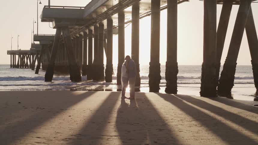 Two people embrace together on the beach underneath a pier