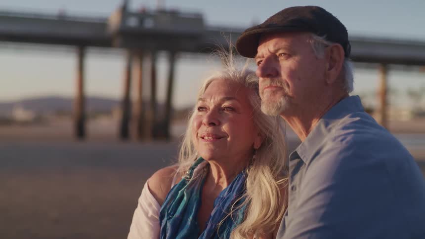 Elderly man with a tweed cap a blue shirt and woman with long grey hair watch sunset on the beach with a pier in the background