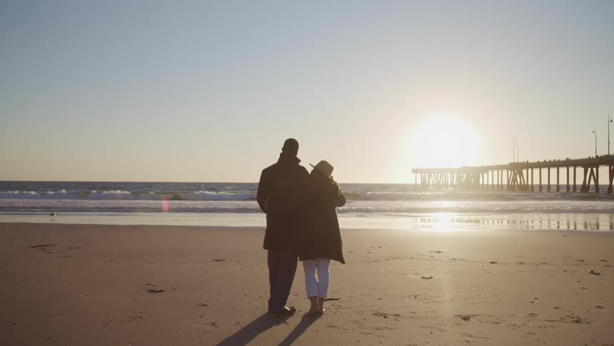 Man with black overcoat and woman with a hat and an overcoat stands and watches sunset on the beach next to the sea and a pier
