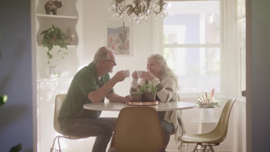 An elderly man and woman talk as they drink coffee in a home