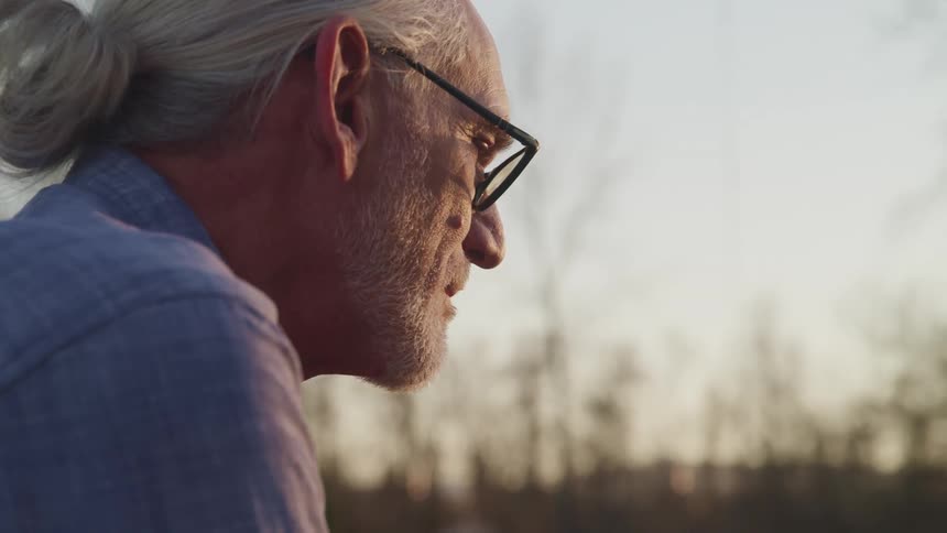 An elderly old man sits outdoors in the evening while lost in thought