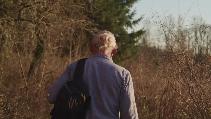 An elderly man uses a hand to shield his eyes from the sun as looks into the distance before he turns and walks through tall brown grass