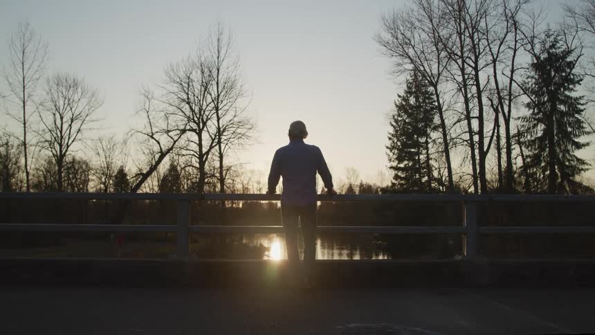 Man stands looking over rail toward river bordered by leafless trees and conifer as sun reflects off water