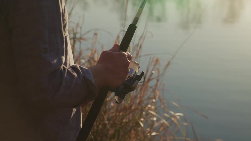 Man fishing with a fishing rod in a lake reels the line with his hands at sunset