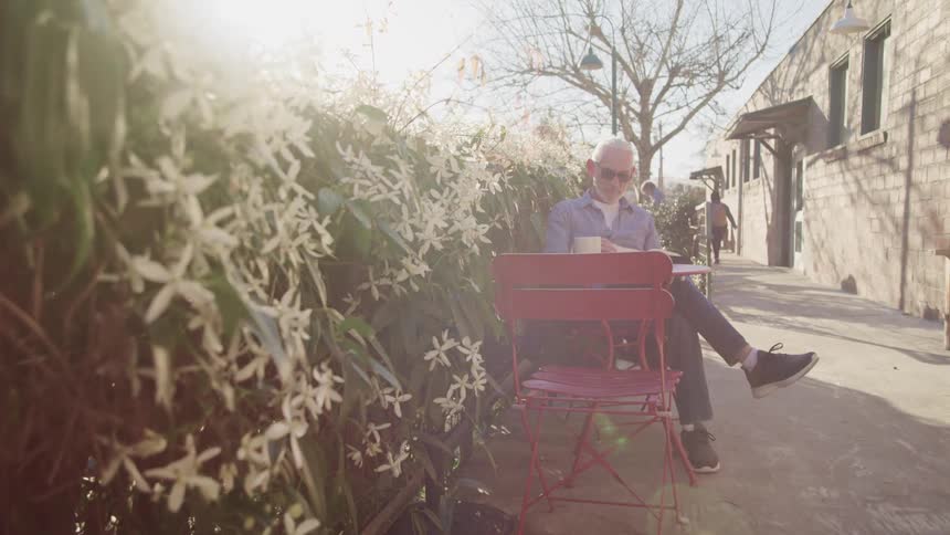Elderly man with long hair a beard and glasses sits outside of a coffee shop at a red table and reads a book