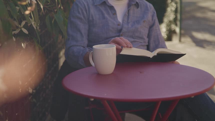A man sitting at a table next to plants and flowers reading a book