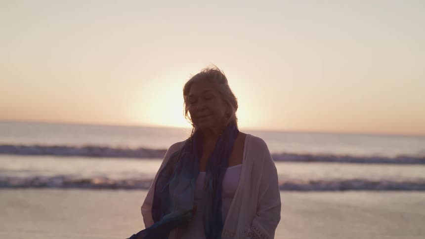 Elderly woman standing on the sand beach next to the sea faces the camera with eye contact and is silhouetted against the sunset