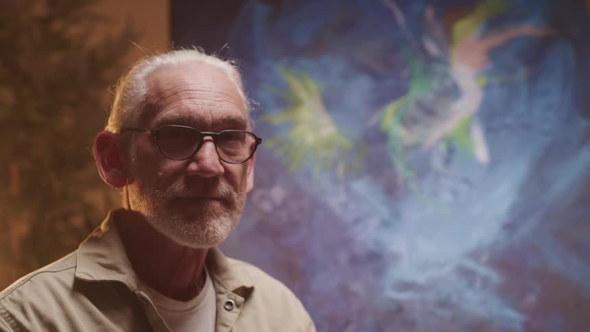 An old man standing in front of a painting raises his head to look into the camera