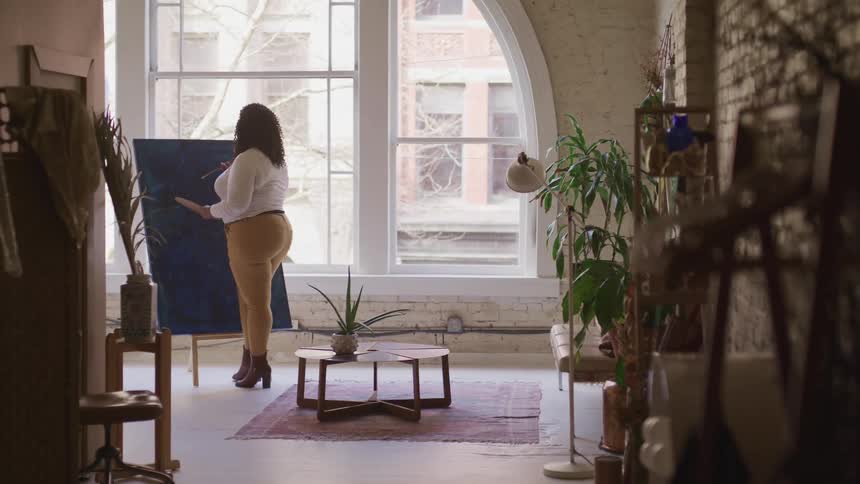 A woman is painting on a canvas on an easel that is placed in front of a large window in a living room