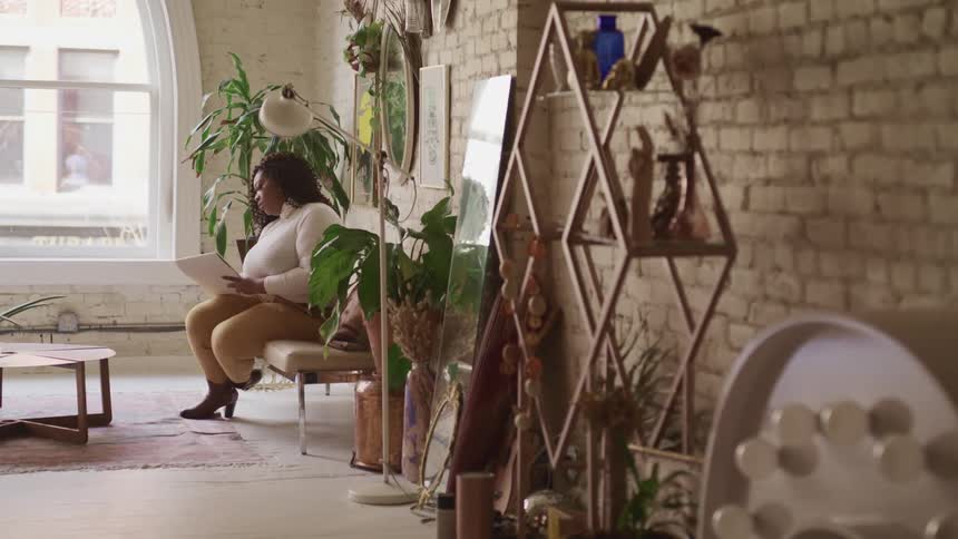 Black woman sits on a chair and reads while in a room with a brick wall