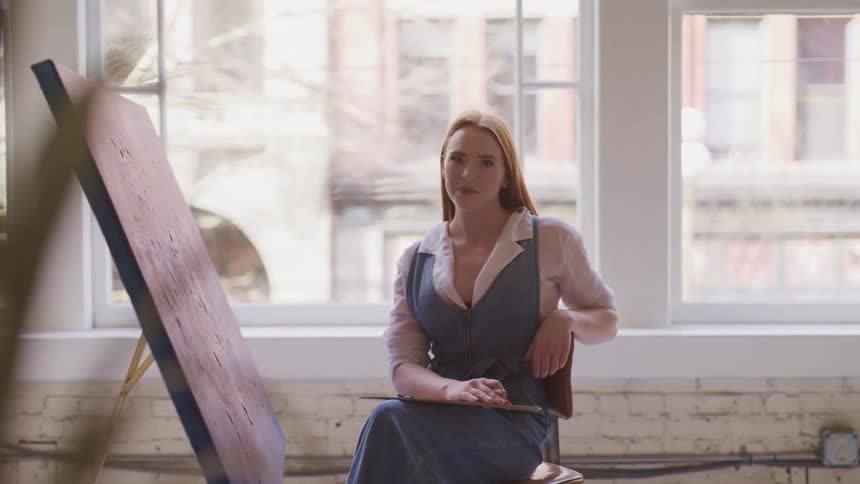 A woman sitting in front of a large canvas turns her head to look into the camera as sunlight enters the large windows behind