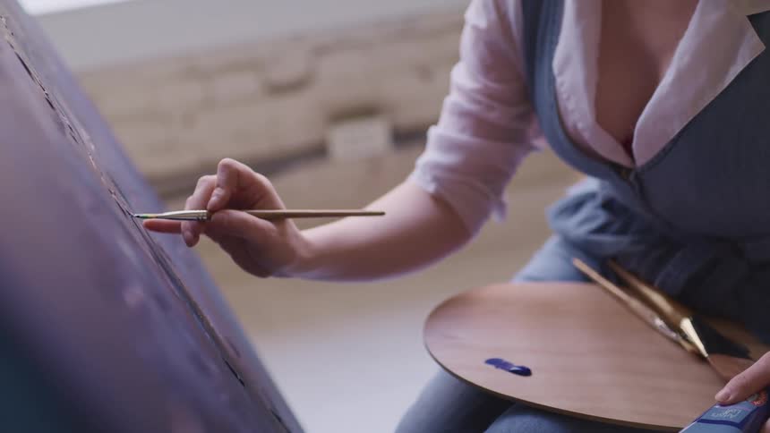 A female artist paints on a canvas while holding a paint palette on her lap