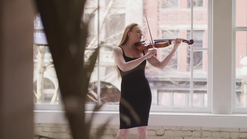 Woman with a black dress holds a violin against her neck and plays by moving a bow