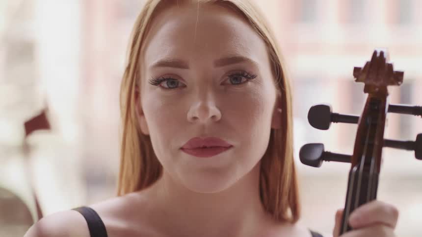 Female violinist with red hair holding a violin faces the camera with eye contact