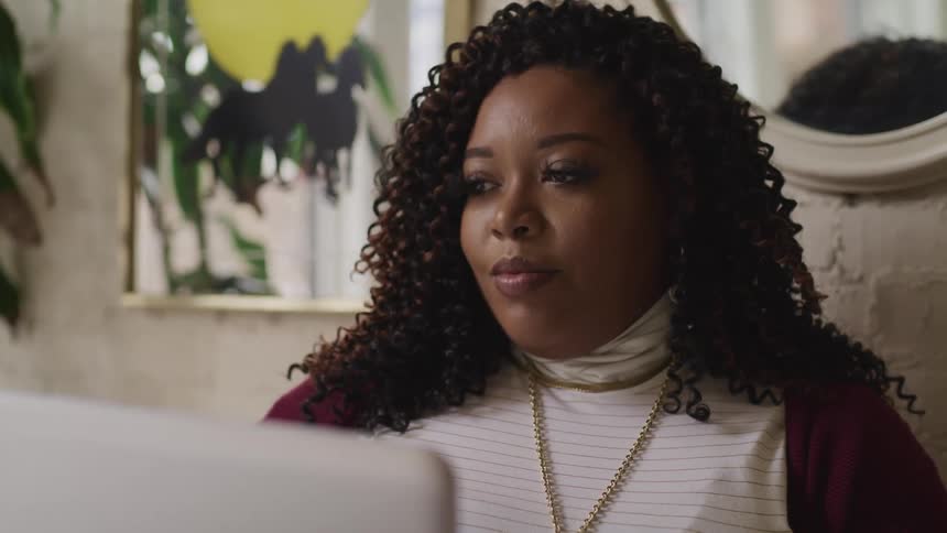 A black woman uses a laptop while sitting next to a plant in a house