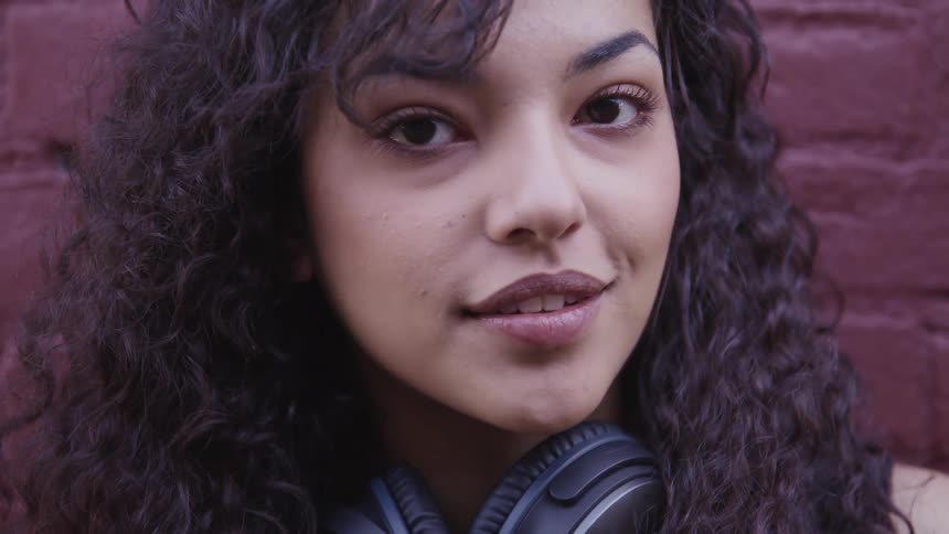A woman stands against a brick wall with headphones around her neck and turns to smile