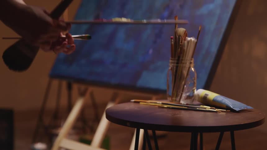 A painter reaches over and picks up a brush an painters easel before walking towards their canvas
