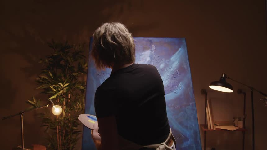A woman in an apron paints on a large canvas with one hand while holding a palette in her other hand