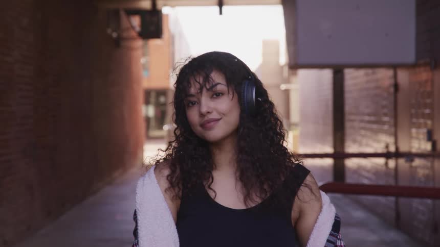 Woman with curly hair and black headphones faces the camera with eye contact and smile in an alley