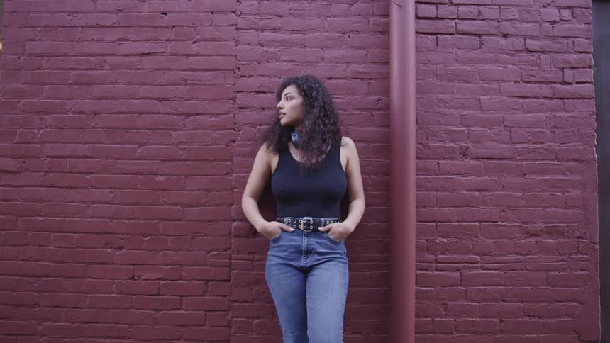 A young hispanic woman with headphones around her neck rests against a building on a sunny day