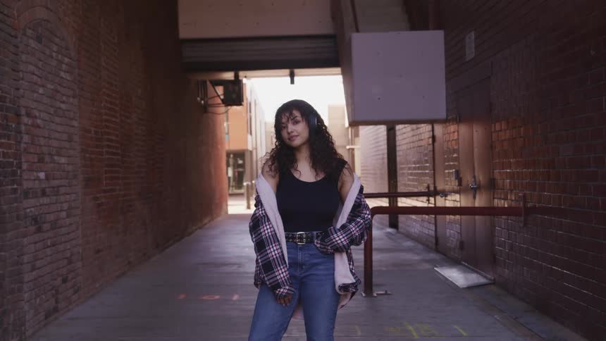 Woman with curly hair and black headphones faces the camera with eye contact and smile in an alley
