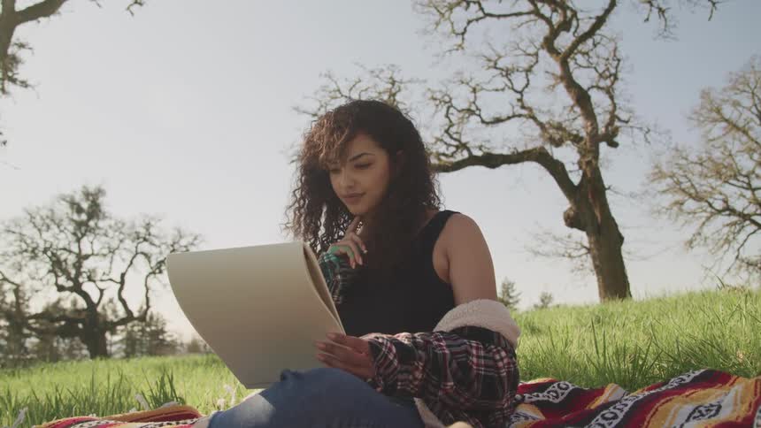 A teenager sitting on a blanket on the grass looks at a pad of paper