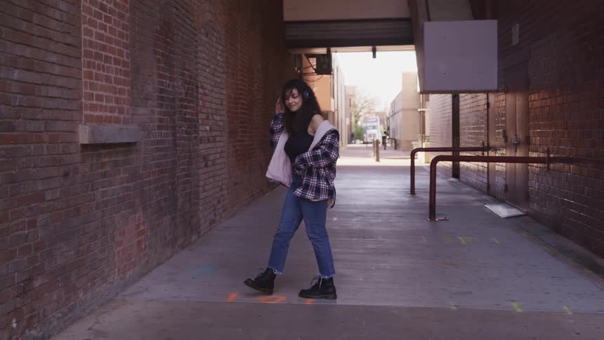 A young woman bobs her head to the music she hears in her headphones as she paces in an alley