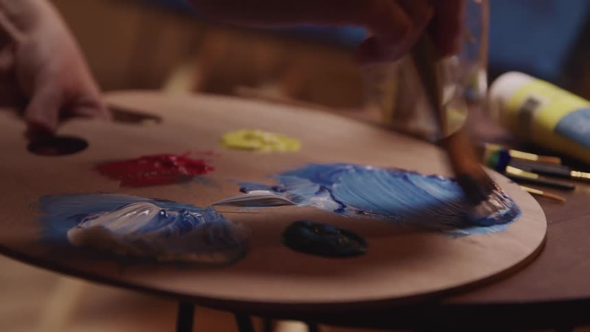 A painter uses a paintbrush to mix paint on a wooden palette