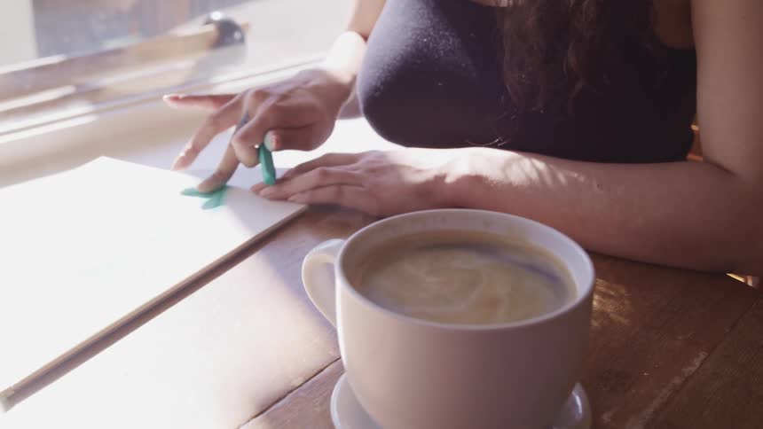 Woman draws a picture using green chalk over white paper next to a big cup of coffee