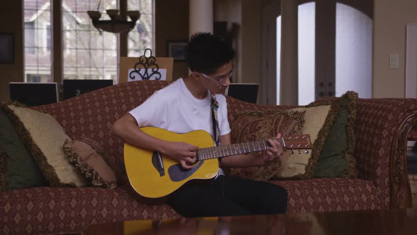 A young male sits on a couch as he strums on an acoustic guitar