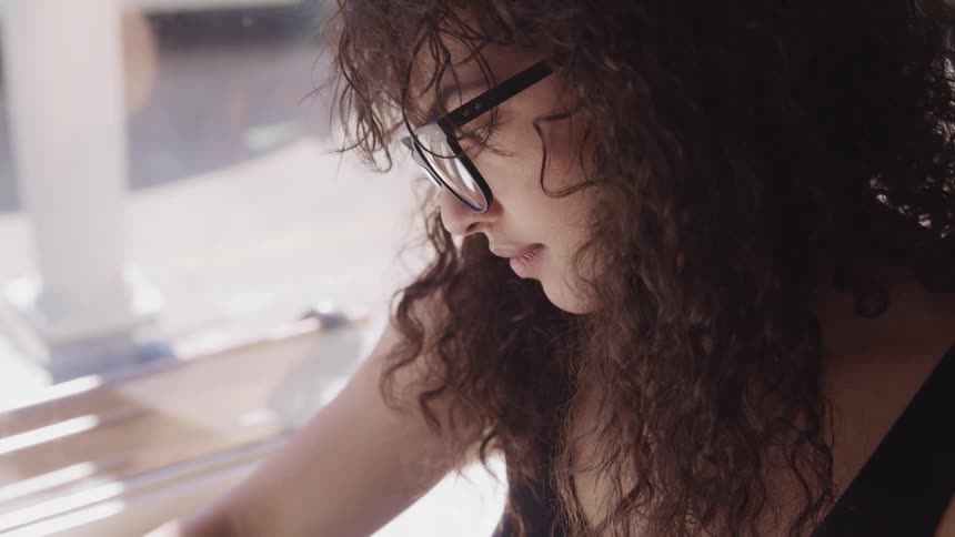 A woman sits inside by a window and looks down as she works on something