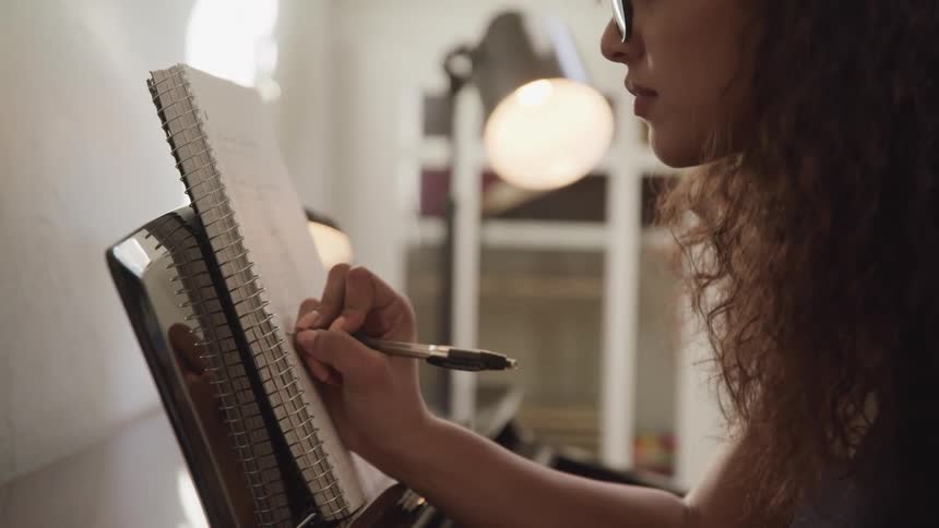 A young woman sits at a piano and writes in a notebook resting in the music rack