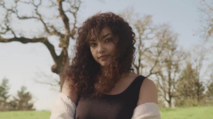 A young woman with dark curly hair poses outside with a sweater worn with the shoulders down