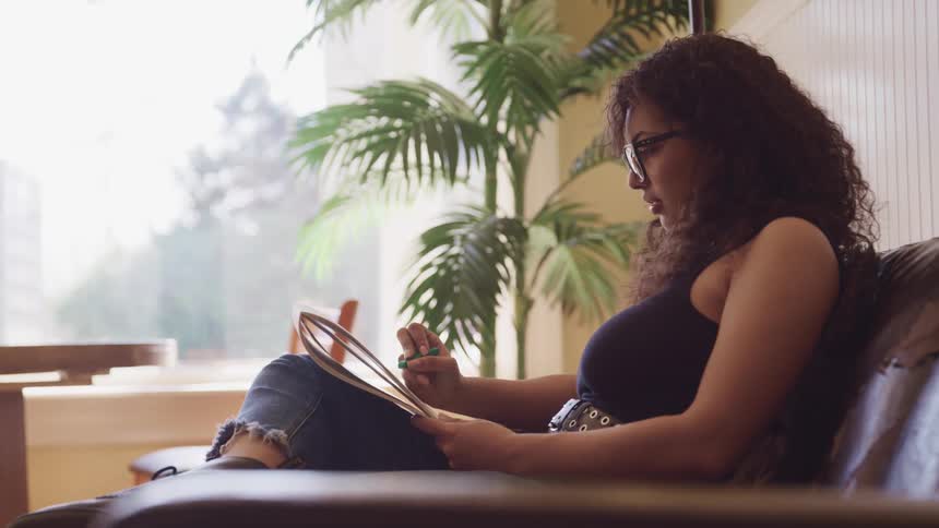 A young woman sits on a leather sofa reading and highlighting the words on a notepad