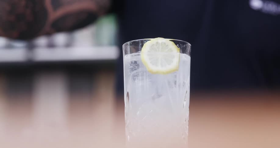 A bartender places a single lemon slice within a glass contain a beverage poured over ice