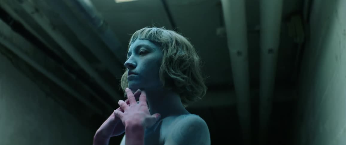 A woman with her face and torso covered in blue powder runs her red powdered hands beneath her chin as she stands in a dim concrete corridor