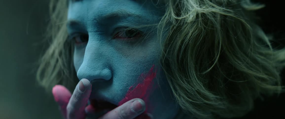Close up of a woman with a painted face applying colored dust to her cheek while looking at the camera