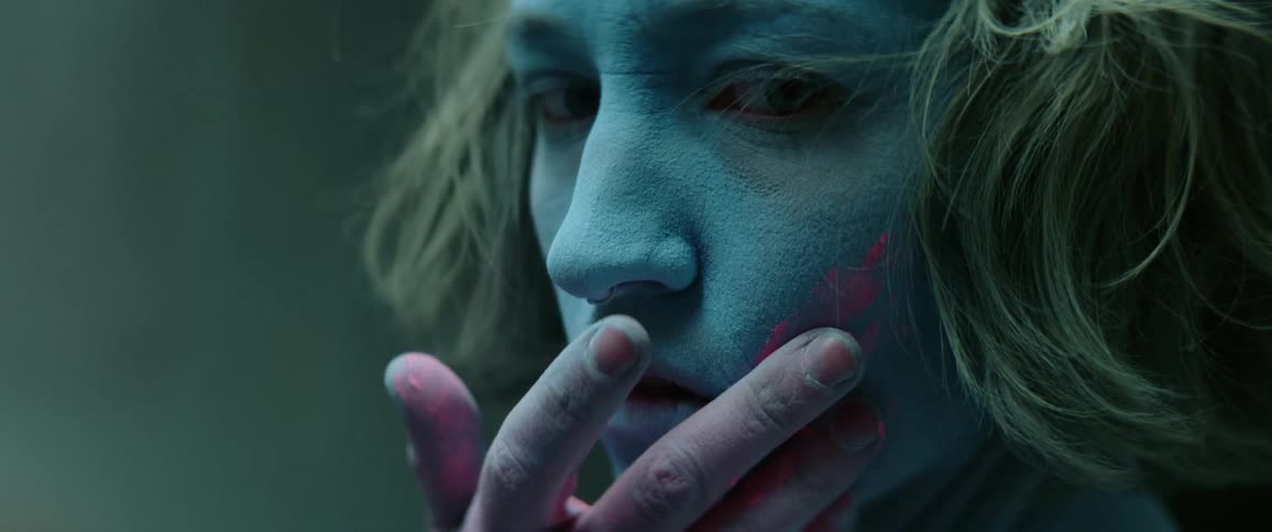 A young woman with blue powder covering her face raises her hand to smear pink powder on her left cheek as she looks into the camera