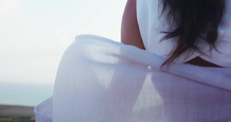 Woman standing on a hill near the ocean wearing a white scarf waving in the wind over her shoulders
