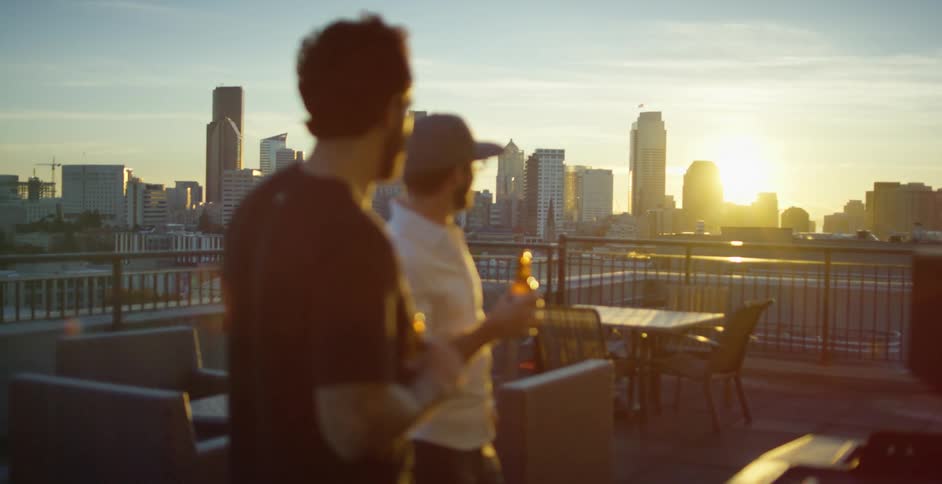 Two men on a roof top patio talk and drink beer and point at the city skyline