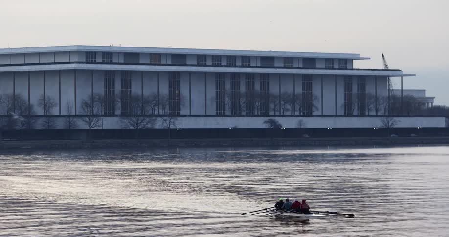 Four people on a row boat sail over the calm waters of a lake near a building