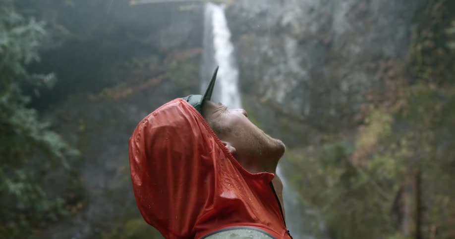 A man standing in front of a waterfall in the rain laughs and opens his mouth towards the sky
