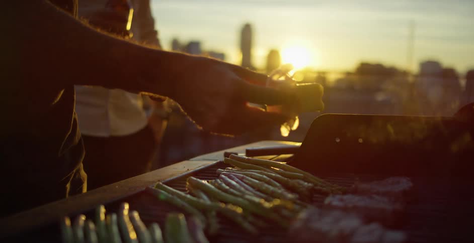 A man rubs butter on asparagus as he grills at home while his friend stands by him and drinks a beer