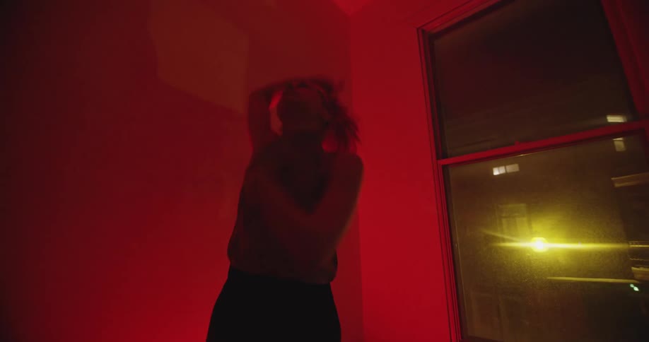 Dancer dances in red light tinted room near window showing city buildings