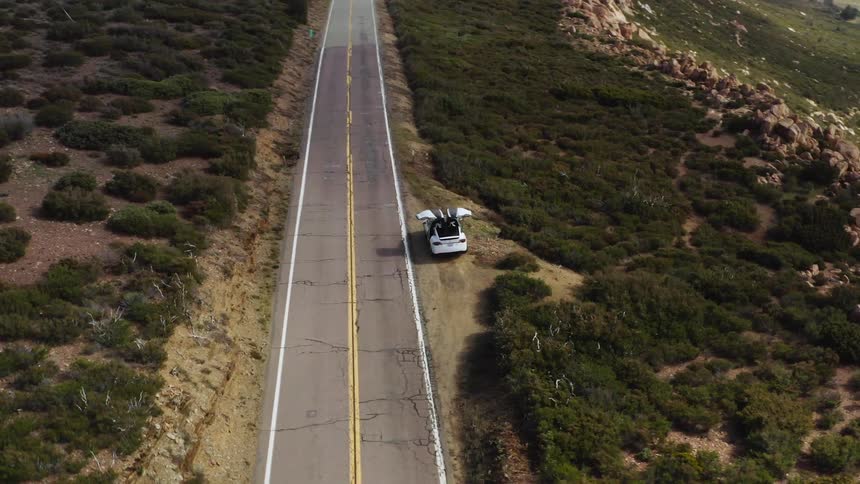 The back doors of a vehicle open vertically as the vehicle rests in the soil on the side of a road running atop a cliff